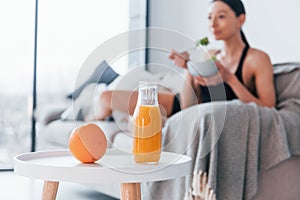 Young woman with slim body shape in sportswear sits on sofa and eats healthy diet food indoors at home