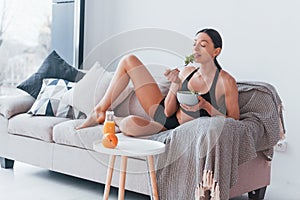 Young woman with slim body shape in sportswear sits on sofa and eats healthy diet food indoors at home