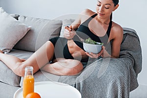 Young woman with slim body shape in sportswear sits on sofa and eats healthy diet food indoors at home