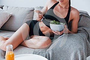 Young woman with slim body shape in sportswear sits on sofa and eats healthy diet food indoors at home