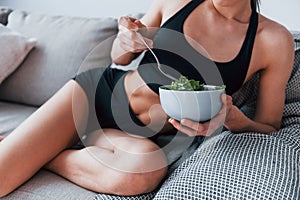 Young woman with slim body shape in sportswear sits on sofa and eats healthy diet food indoors at home