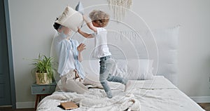 Young woman and small boy enjoying pillow fight on bed in apartment