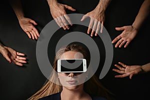 Young woman with smartphone covering her eyes surrouded by people`s hands on black background, top view