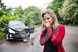 A young woman with smartphone by the damaged car after a car accident, making a phone call.