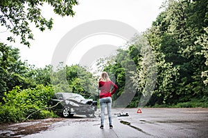 A young woman with smartphone by the damaged car after a car accident, making a phone call.