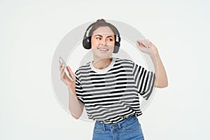 Young woman with smartphone listening to music, dancing to her favourite song in headphones, posing against white