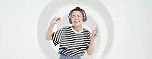 Young woman with smartphone listening to music, dancing to her favourite song in headphones, posing against white