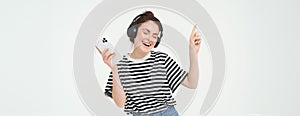 Young woman with smartphone listening to music, dancing to her favourite song in headphones, posing against white