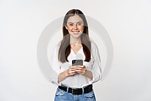 Young woman with smartphone, smiling and looking at camera, using mobile phone app, cellular technology and online