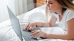 Young woman smile typing on laptop keyboard while lying in bed at home. Happy teen girl communicates online working