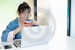 Young woman smiling holding the cup of coffee and looking at laptop screen.