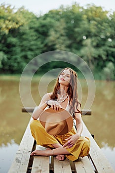 A young woman smiling in an image of a hippie and eco-clothes sitting outdoors on a bridge by a lake wearing a hat and