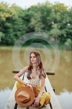 A young woman smiling in an image of a hippie and eco-clothes sitting outdoors on a bridge by a lake wearing a hat and
