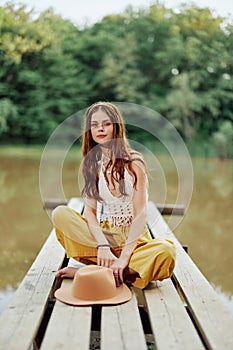 A young woman smiling in an image of a hippie and eco-clothes sitting outdoors on a bridge by a lake wearing a hat and