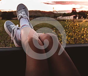 Young woman with sneakers with feet propped on the car window at sunset