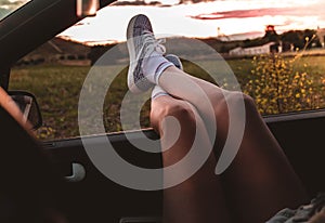 Young woman with sneakers with feet propped on the car window at sunset