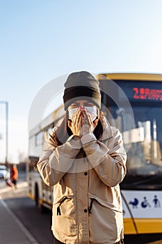 Young woman sneezes on a bus station and covers her face with hands. Girl wears a white medical mask standing near a bus at a