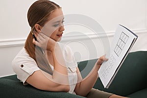 Young woman solving sudoku puzzle on sofa indoors