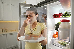 Young woman with spoiled sausage near refrigerator in kitchen