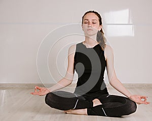 Young woman in sports clothes meditating in lotus position while doing yoga at home