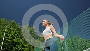 Young woman in sportswear does jump exercise at sports ground with skipping rope