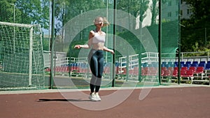 Young woman in sportswear does jump exercise at sports ground with skipping rope