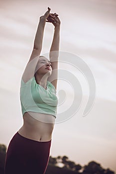 Young woman in sportswear exercises and stretches hands up on background of cloudy sky, girl engaged in sport outdoors, concept