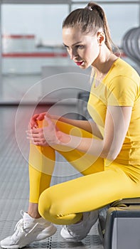 Young woman in sportswear having pain in her knee while training in gym, Girl sitting on a floor touching her knee in