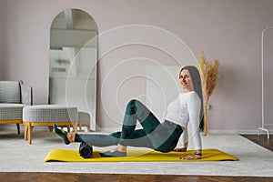 Young woman in sportswear performs a myofascial massage of the lower leg muscles using a roller or roll on a gym mat at home.