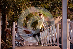 Young woman in sportswear is resting after workout, walking along wooden stairs in the rays of sun