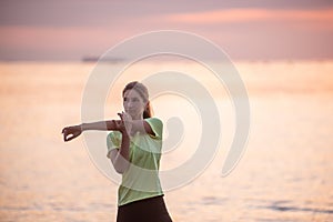 Young woman in sportswear is warming up early in morning on seashore at sunrise. Fitness