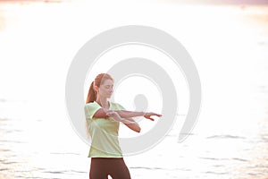 Young woman in sportswear is warming up early in morning on seashore at sunrise. Fitness