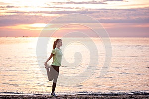 Young woman in sportswear is warming up early in morning on seashore at sunrise. Fitness