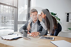 Young woman stand besides man and whispering on his ear. Guy listen to her and look at model. They are in meeting room