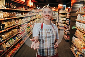 Young woman stand in line between two pasta shelfs in grocery store. She reach hand to camera and smile. Model posing.