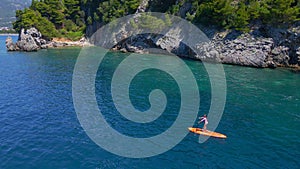 Young woman on a stand up paddle board SUP rawing among beatyful rocks. Clear blue sea water surrounds her. Aerial