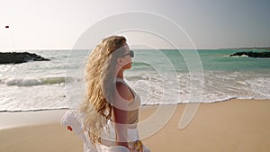 Young woman standing, curly long hair blowing in wind at sandy beach, sea surf. Attractive girl in sunglasses posing