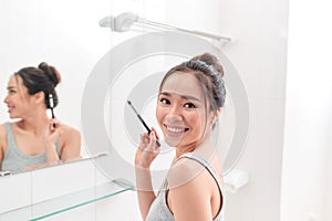 A young woman is standing in front of the bathroom mirror and putting on makeup