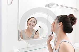 A young woman is standing in front of the bathroom mirror and putting on makeup