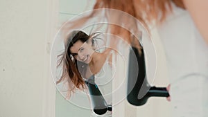Young woman standing in front of mirror in pajamas. Girl using hair dryer and sing, wind blowing the hair. Slow motion.