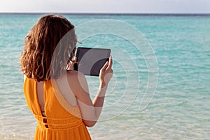 Young woman standing in front of the sea and using her tablet during sunset