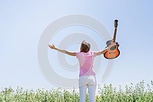 Young woman standing in grass fiel raising her hand to the sky, holding guitar