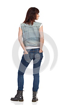 Young Woman Is Standing, Holding Hands In Pocket And Looking Away. Rear View.
