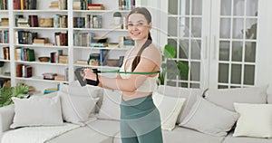 Young woman is standing in the living room getting ready to do exercises with an expander, looking at the video camera