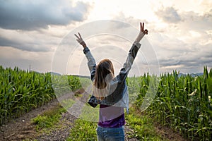 Young woman standing in the middle of green summer corn field with her arms raised high in victorious gesture
