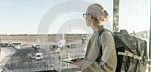 Young woman standing near airport gates window holding cellphone in her hands, wearing travel backpack and walking to