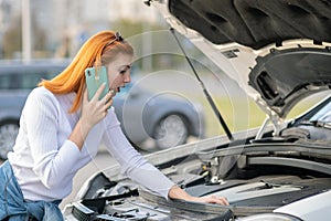 Young woman standing near broken car with popped hood talking on her mobile phone while waiting for help