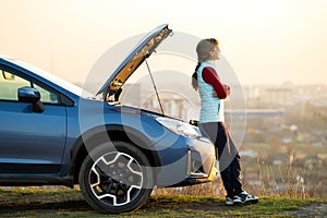 Young woman standing near broken down car with popped up hood having trouble with her vehicle. Female driver waiting for help