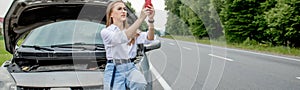 Young woman standing near broken down car with popped up hood having trouble with her vehicle. Waiting for help tow truck or