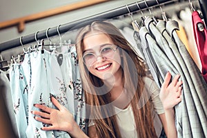 Young woman standing near clothes rack on color background